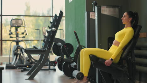 Strong-hispanic-brunette-sportswoman-doing-legs-exercise-on-sport-simulator-in-fitness-club.-In-yellow-sportswear.-Athletic-woman-performs-an-extension-leg-exercise-in-the-simulator